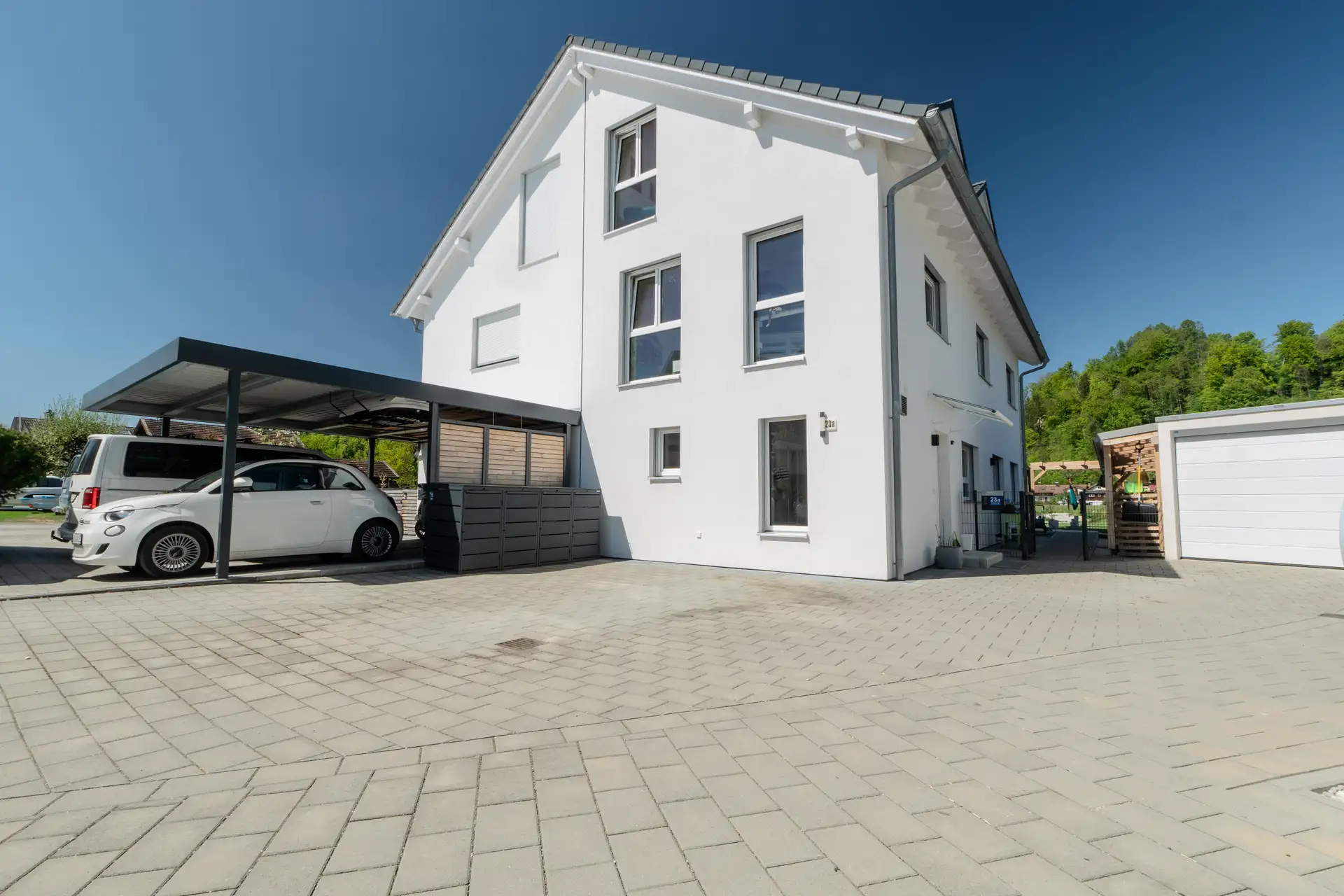 Modernes weißes Doppelhaus mit Carport und gepflasterter Einfahrt in Wolfratshausen, im Hintergrund bewaldeter Hang