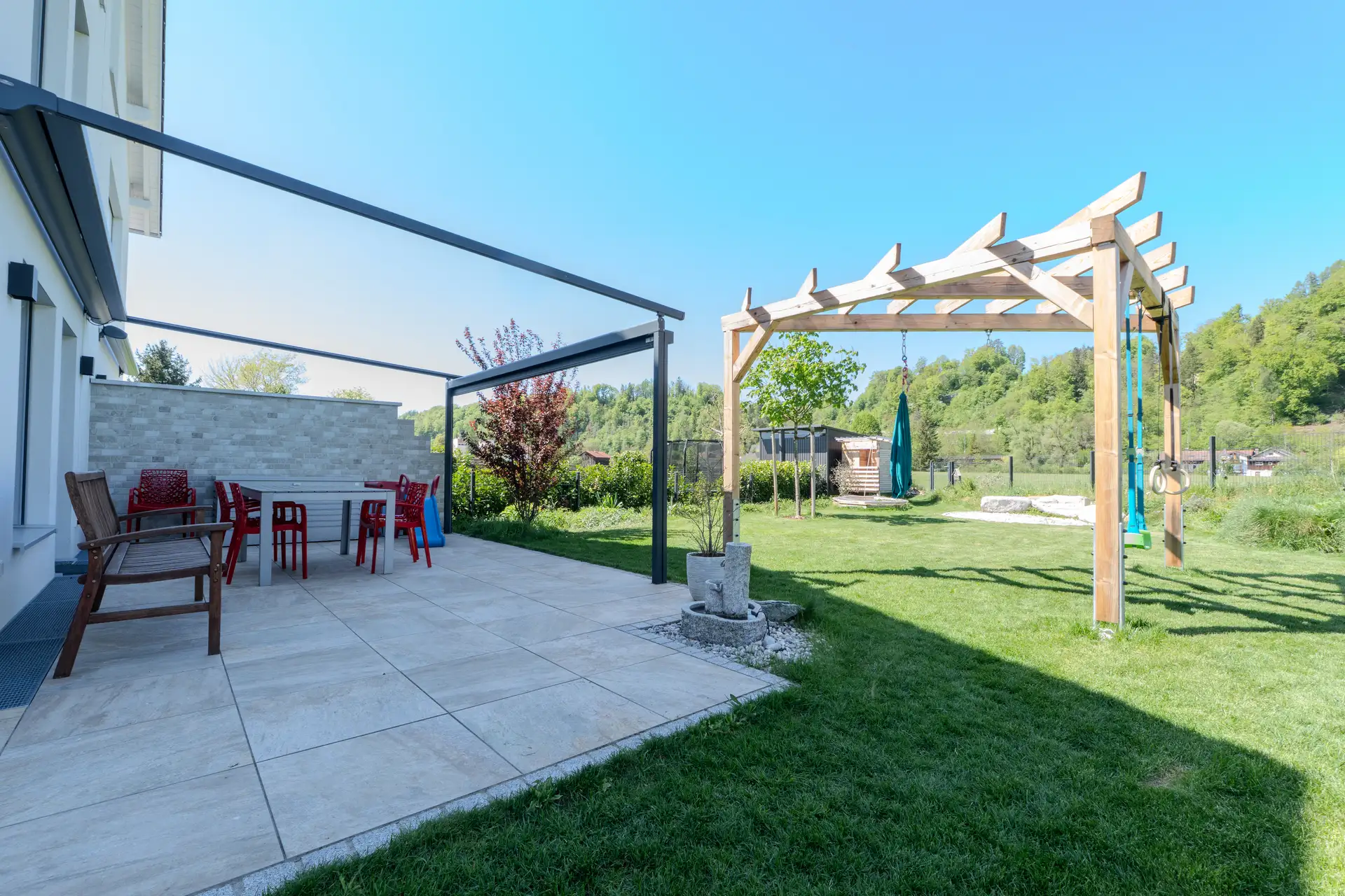 Terrasse mit Pergola und Garten eines Einfamilienhauses in Wolfratshausen, dahinter der bewaldete Hang
